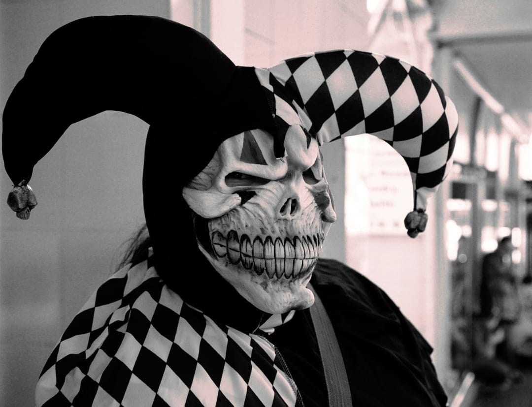 Jester wearing a skeletal mask and a two-pointed bell hat in a grayscale portrait with a blurred background.
