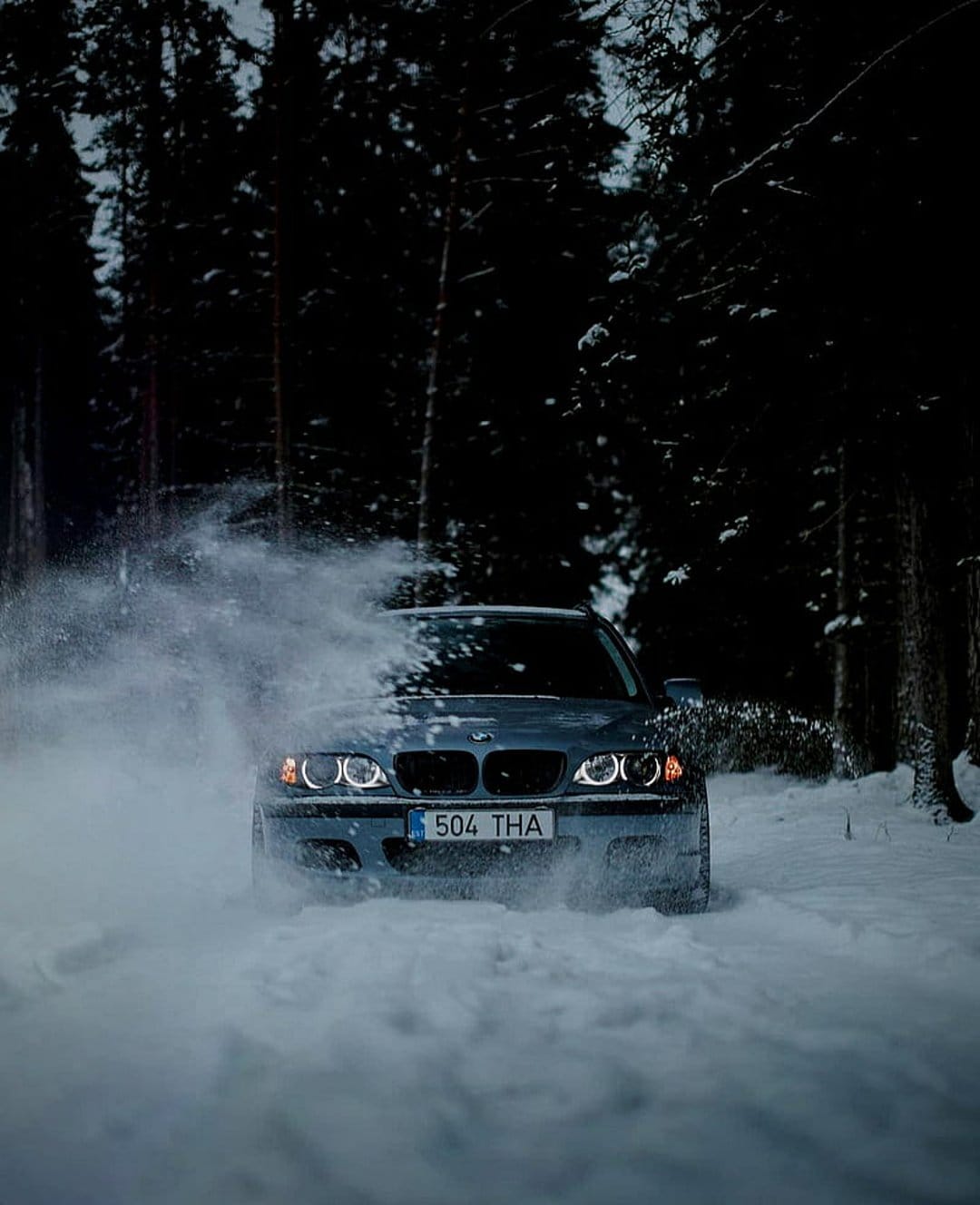 BMW sedan drifting on a snowy forest road at night with glowing headlights and high-speed powder spray.