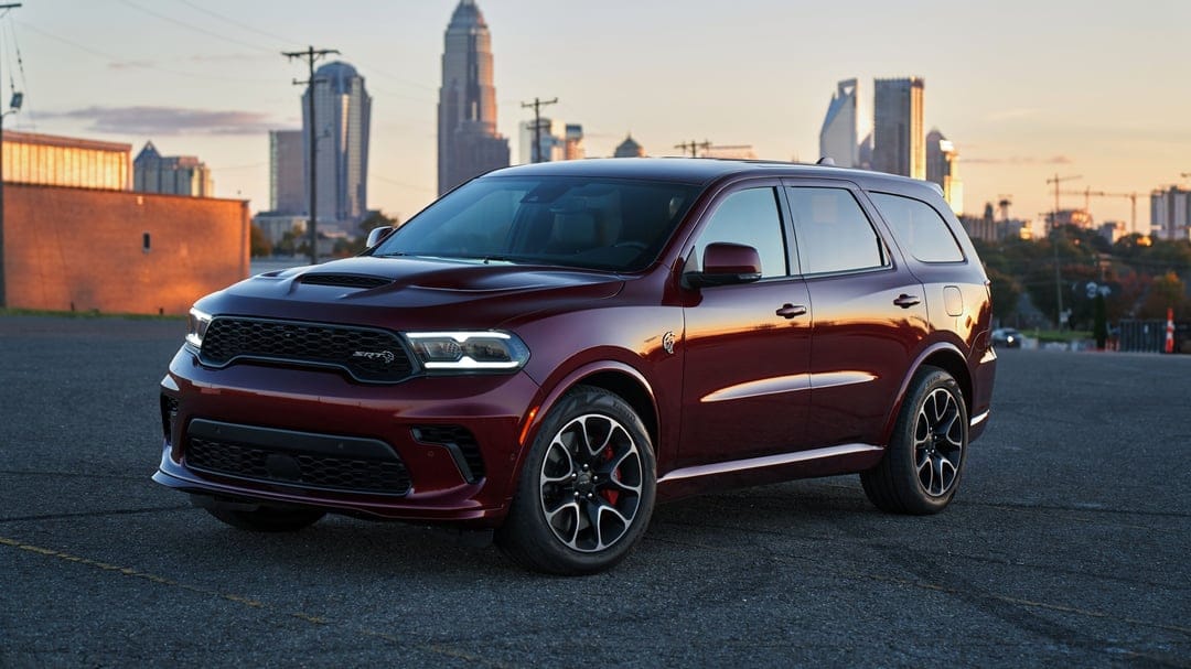 Maroon Dodge Durango SRT Hellcat parked on asphalt with a glowing city skyline in the background at sunset.