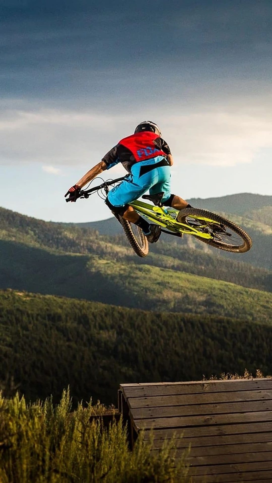 Mountain biker in mid-air above a wooden ramp with rolling green hills and a dramatic sky in the background.