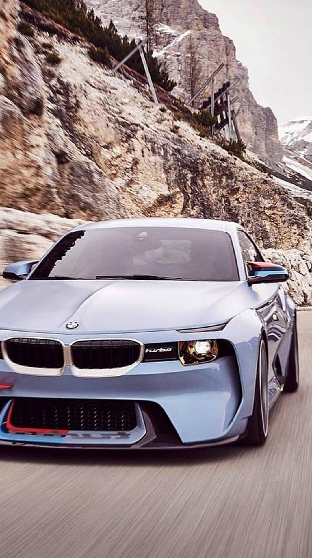 Blue BMW Turbo concept car driving on a winding mountain road with rocky slopes and snowy peaks in the background.