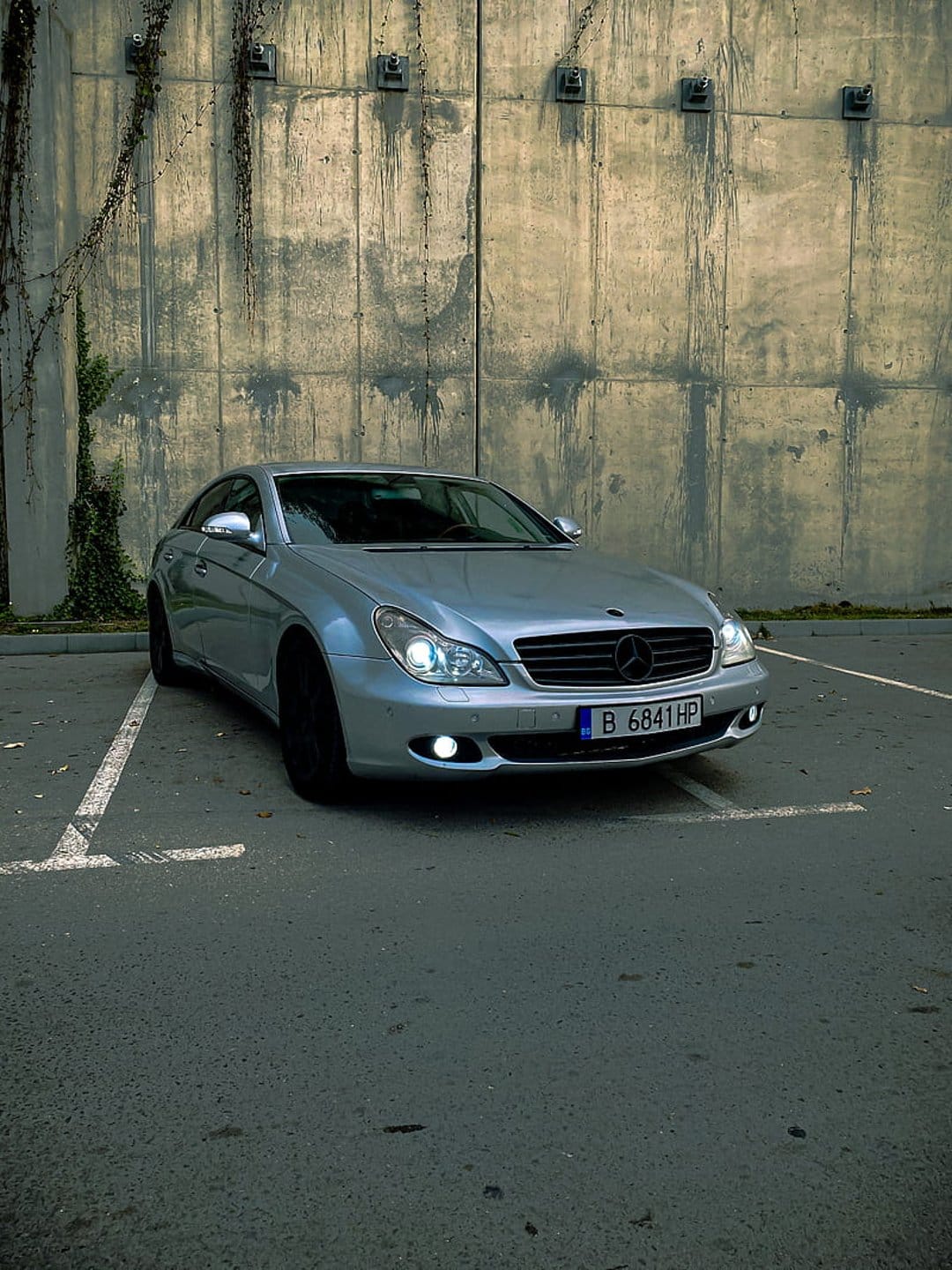 Silver Mercedes CLS sedan with glowing headlights parked before a concrete wall and ivy on an asphalt lot.