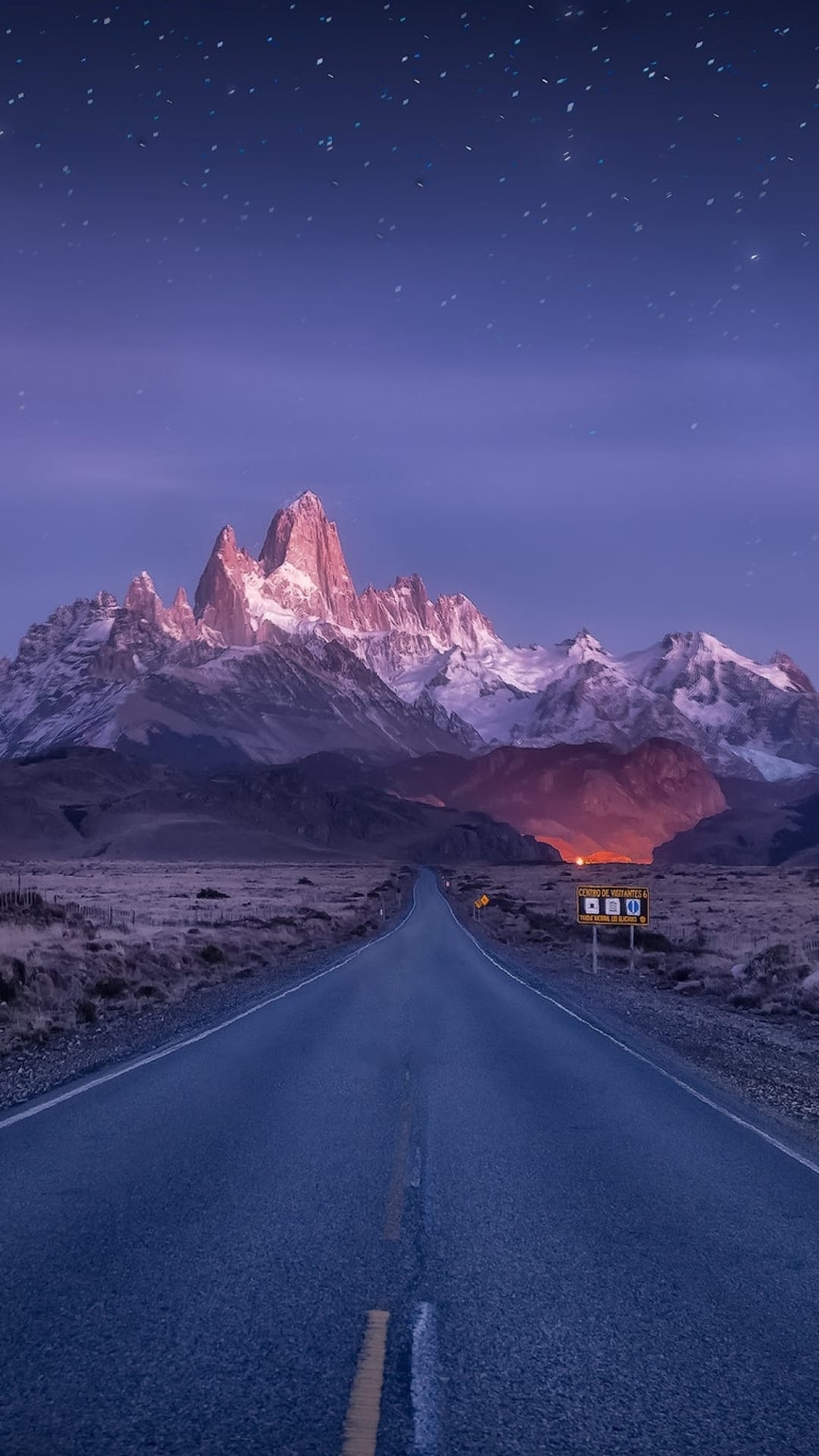 Straight asphalt road extending toward snow-capped granite mountains under a dense, starry twilight sky.