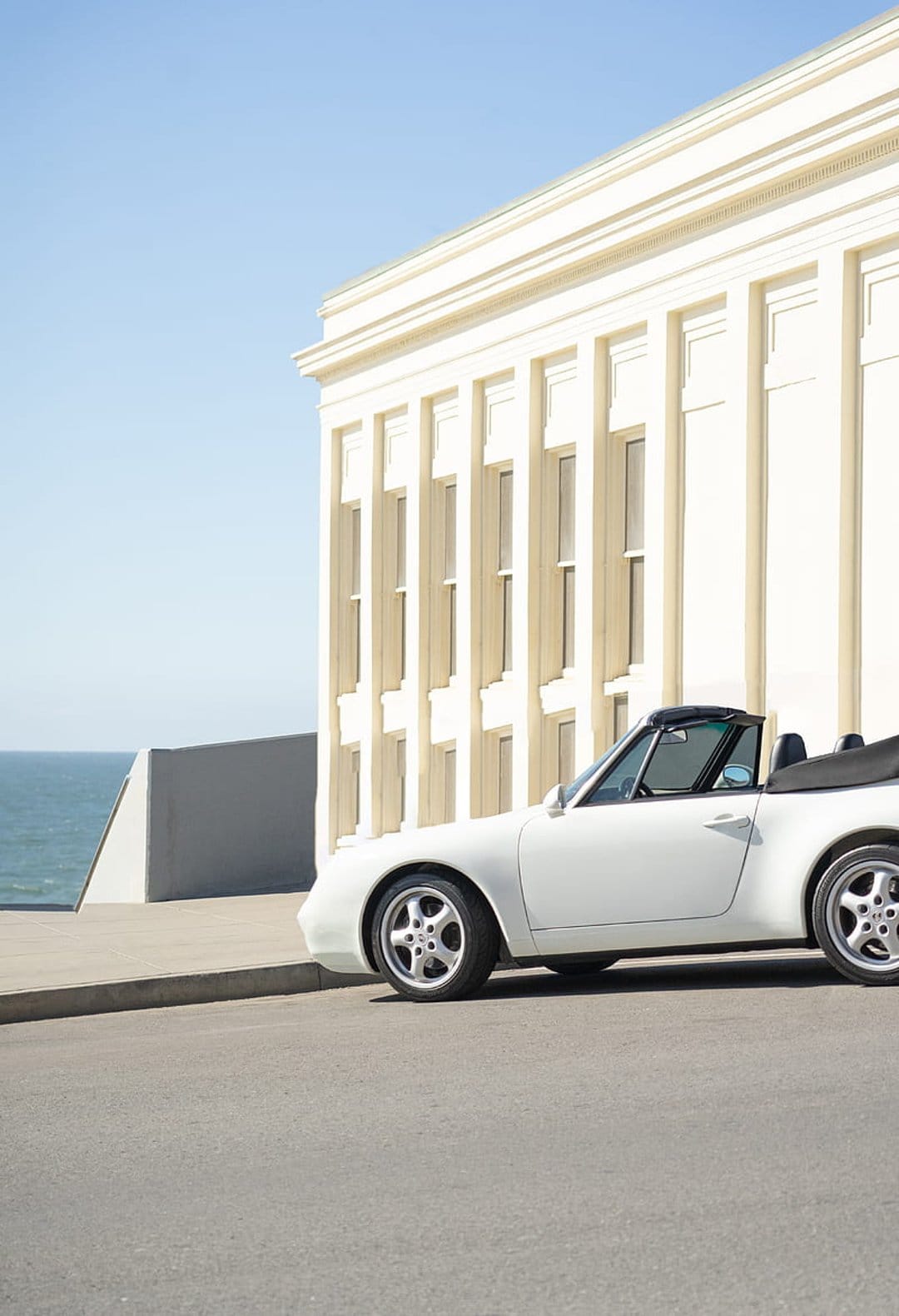 White convertible car parked on a road beside a cream minimalist building with the blue ocean in the background.