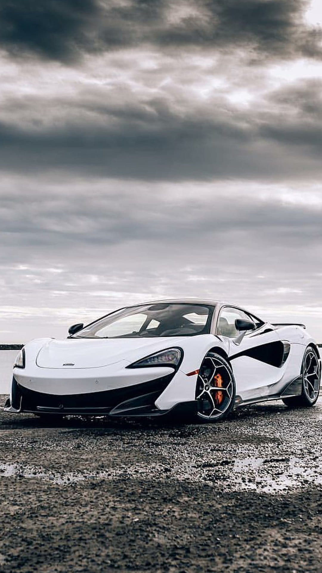 White McLaren sports car with orange calipers parked on a wet rocky shore beside a lake under heavy grey clouds.