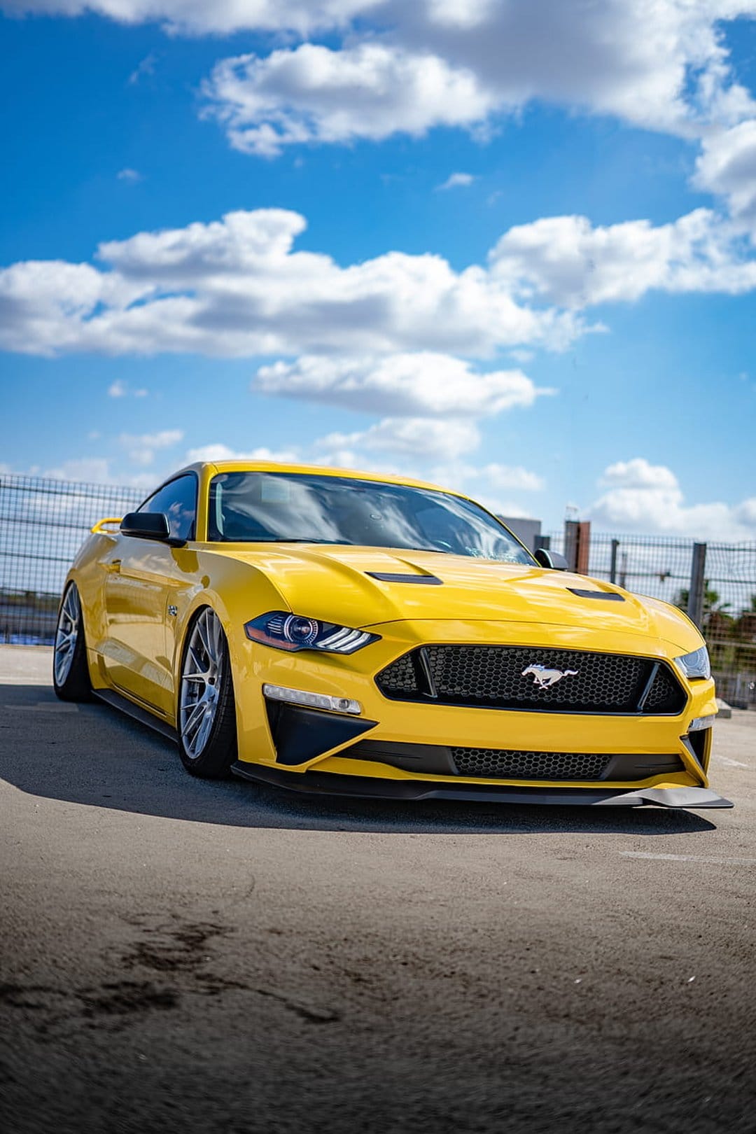Yellow Ford Mustang convertible with its top down parked on asphalt under a sunny blue sky with white clouds.