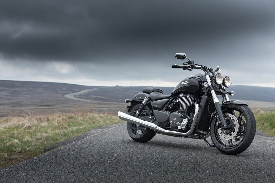 Black Triumph motorcycle parked on a scenic road under a heavy dark storm sky with distant rolling hills.