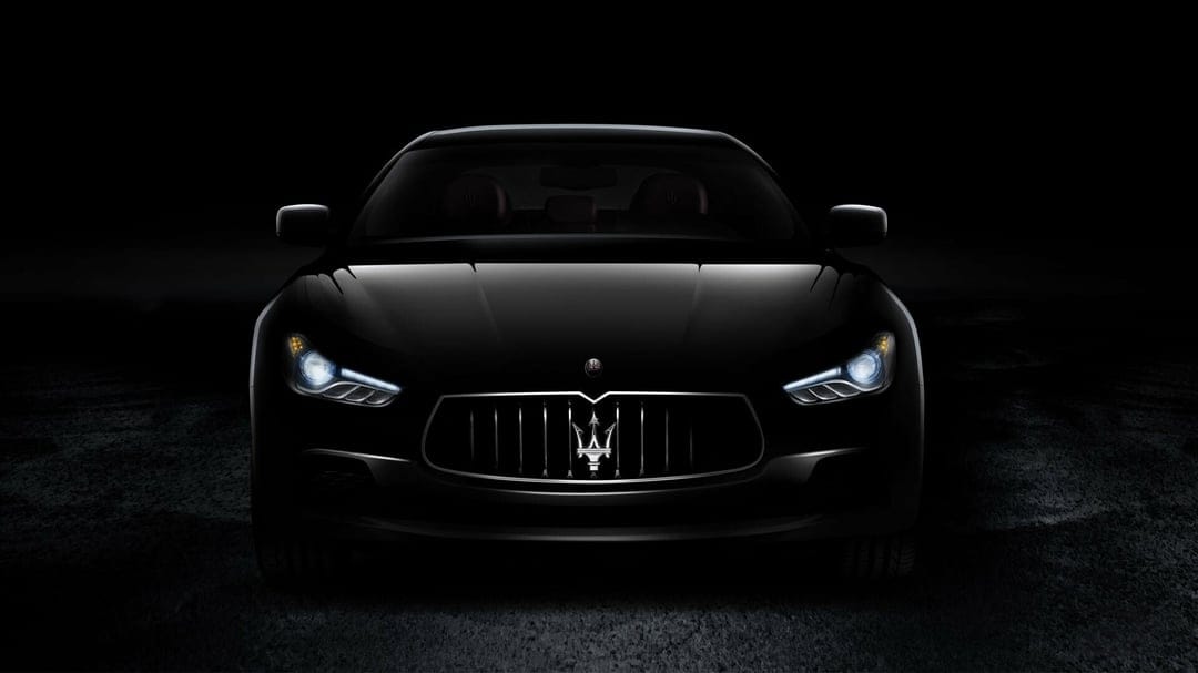 Front view of a black Maserati Ghibli with glowing headlights and trident logo in a dark, moody environment.