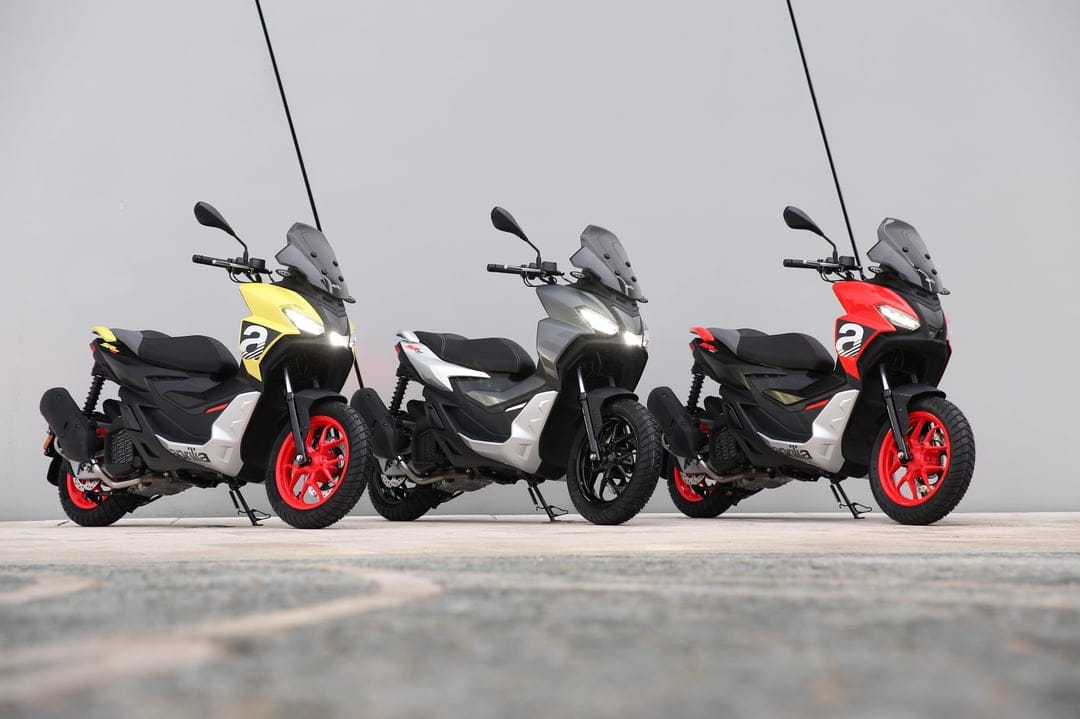 Three Aprilia SR GT scooters in yellow, silver, and red with headlights on against a grey wall background.