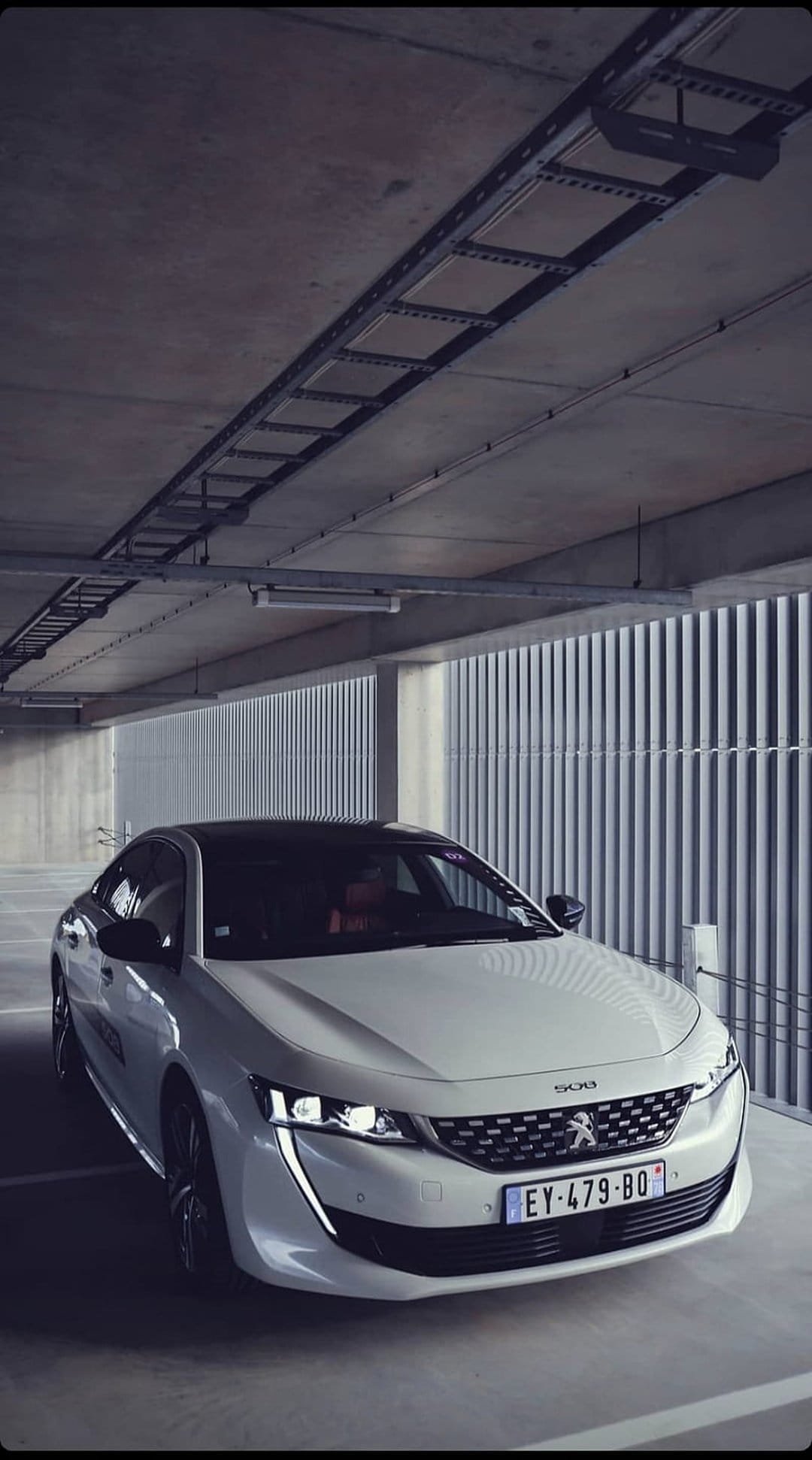 White Peugeot 508 sedan parked in a concrete underground garage with glowing LED headlights and metal textures.
