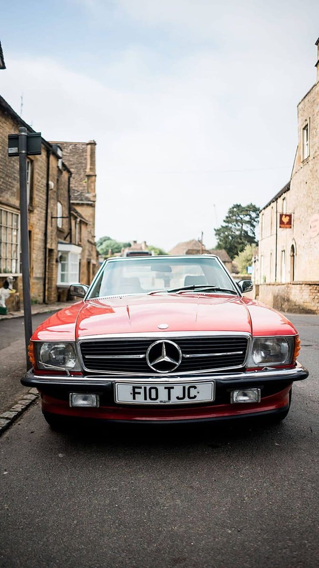 Red Mercedes SL roadster parked on a cobblestone street beside historic stone cottages in an English village.