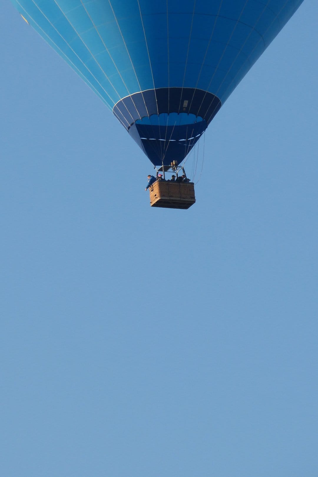 Blue hot air balloon with a wicker basket floats upward through a vast, cloudless azure sky on a clear day.