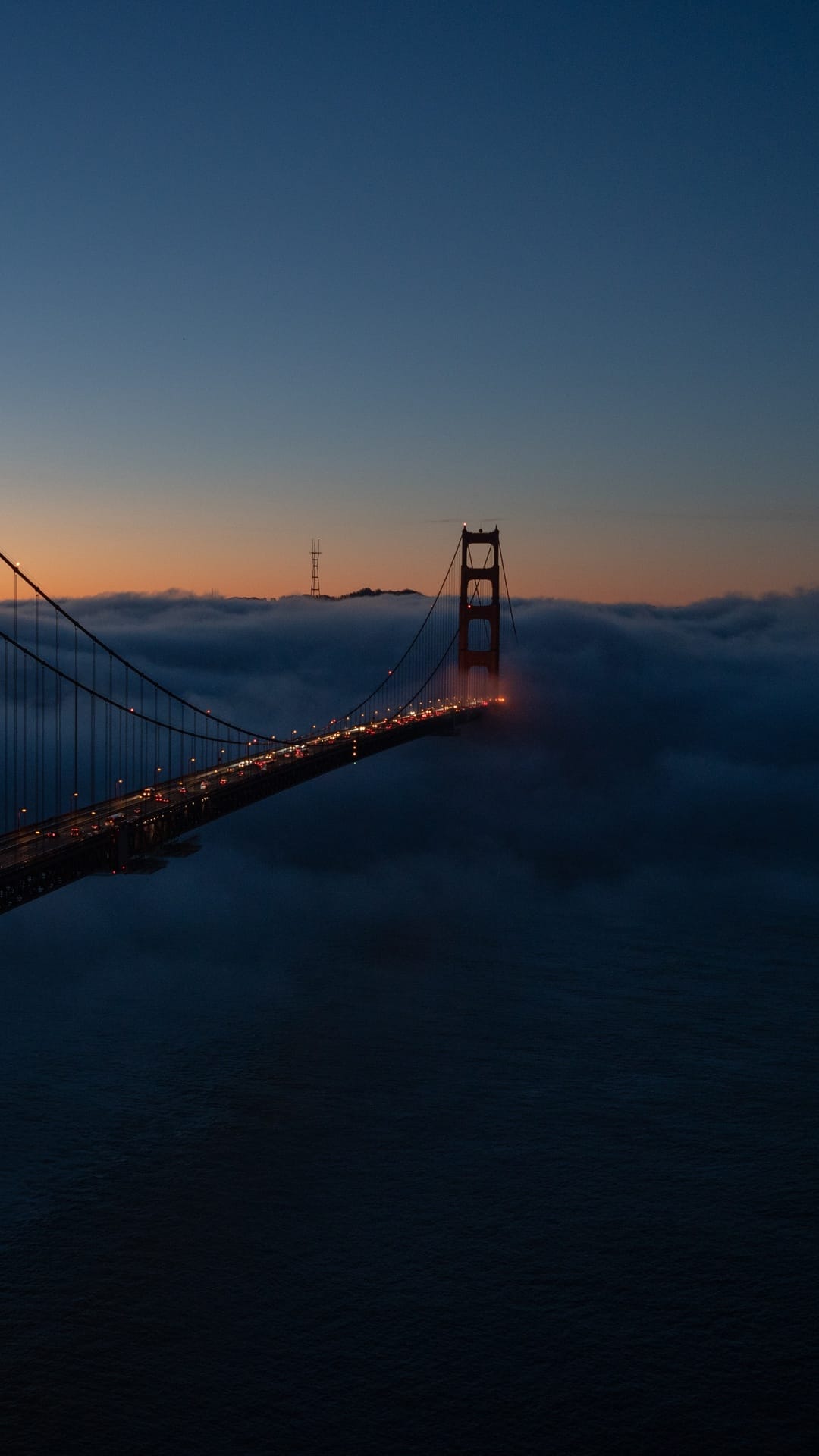 Golden Gate Bridge towers rise from a thick layer of fog at twilight under a gradient orange and blue sky.