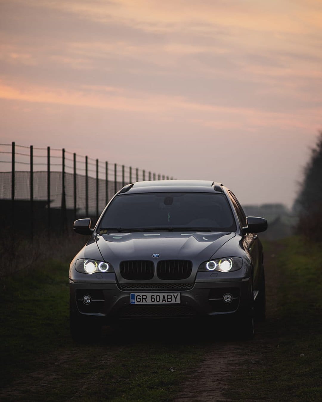 BMW X6 SUV with illuminated headlights parked on a dirt road against a pastel sunset sky and rural trees.
