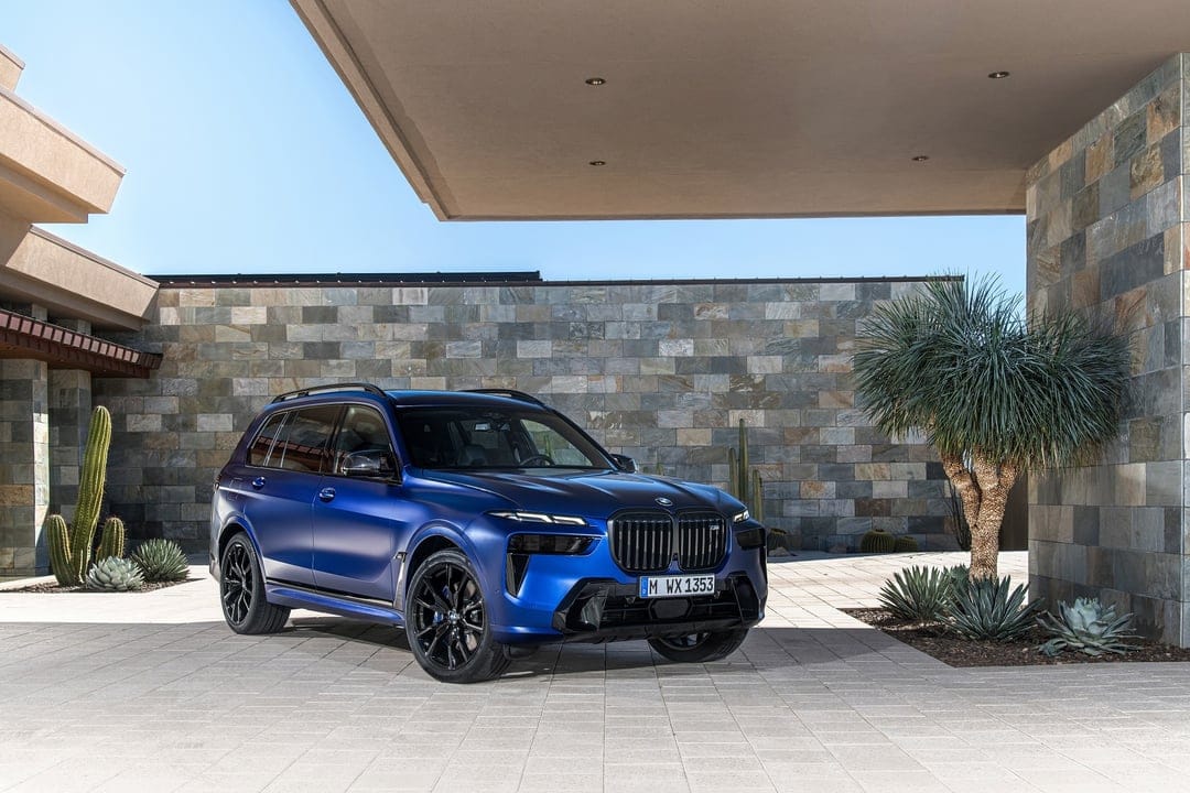 Blue BMW X7 M60i SUV parked on a paved driveway in front of a modern stone wall with desert cacti and palms.