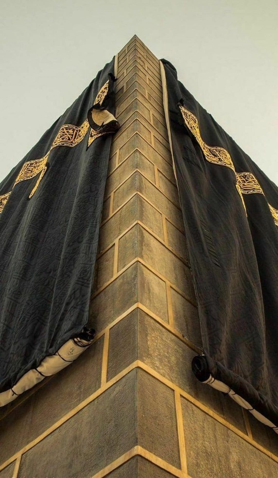 Kaaba corner featuring black silk Kiswah fabric with ornate gold embroidery over textured grey stone masonry.