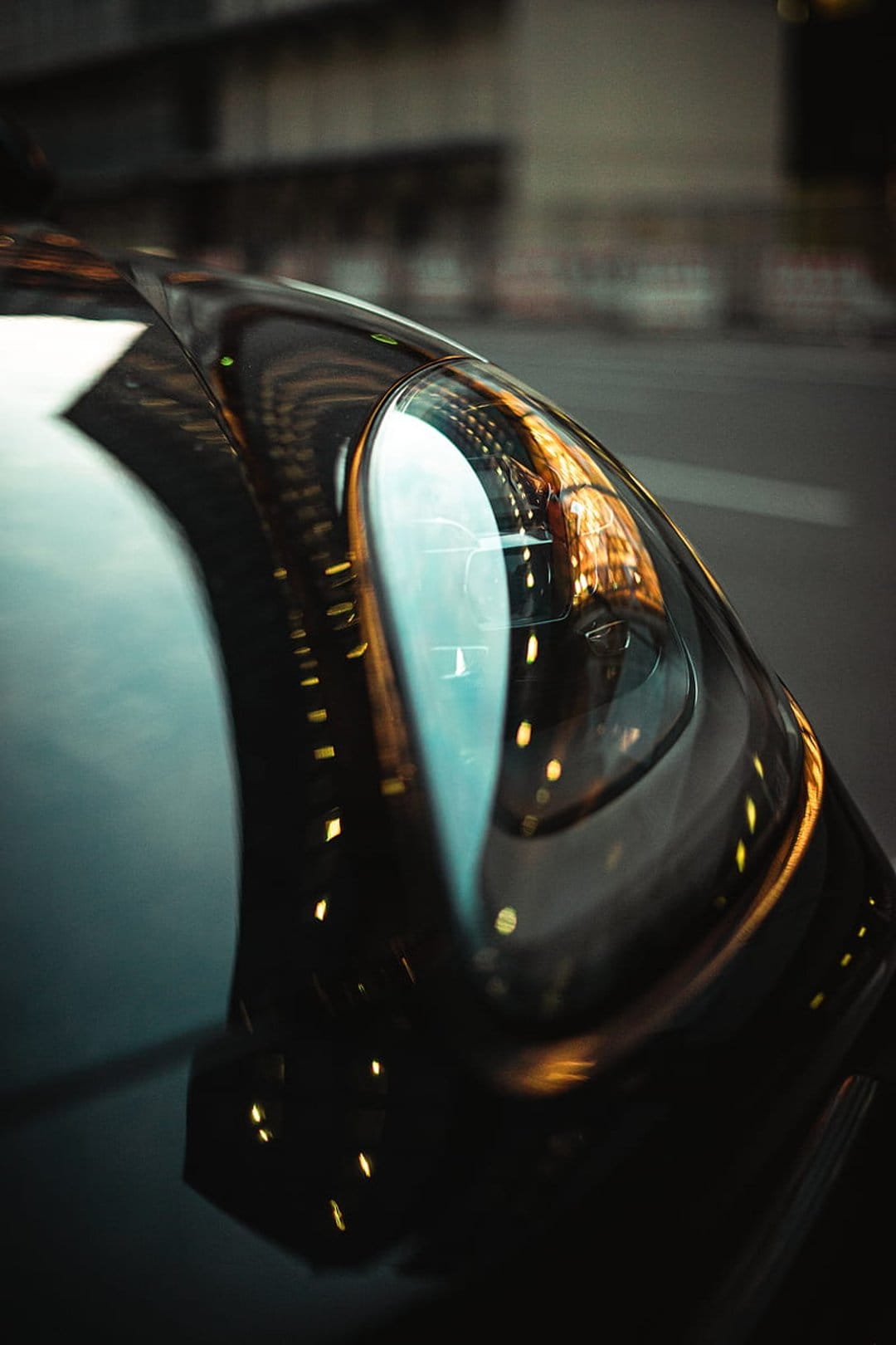 Sleek black car headlight reflecting colorful urban neon lights and bokeh against a dark metallic exterior.