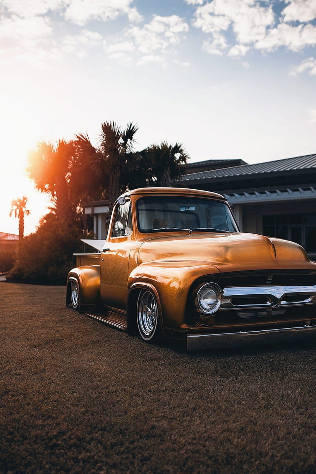 Golden vintage pickup truck with chrome wheels parked near palm trees under a warm sunset sky in an urban setting.