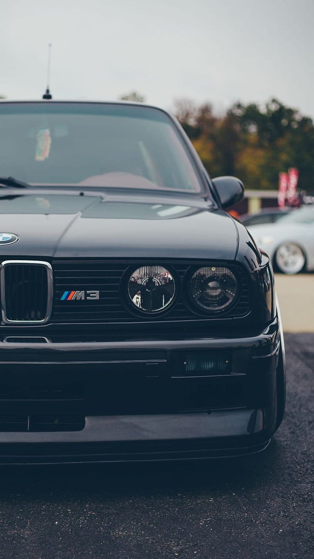 Dark gray BMW E30 M3 front grille and circular headlights with the M3 logo badge on a vintage car chassis.