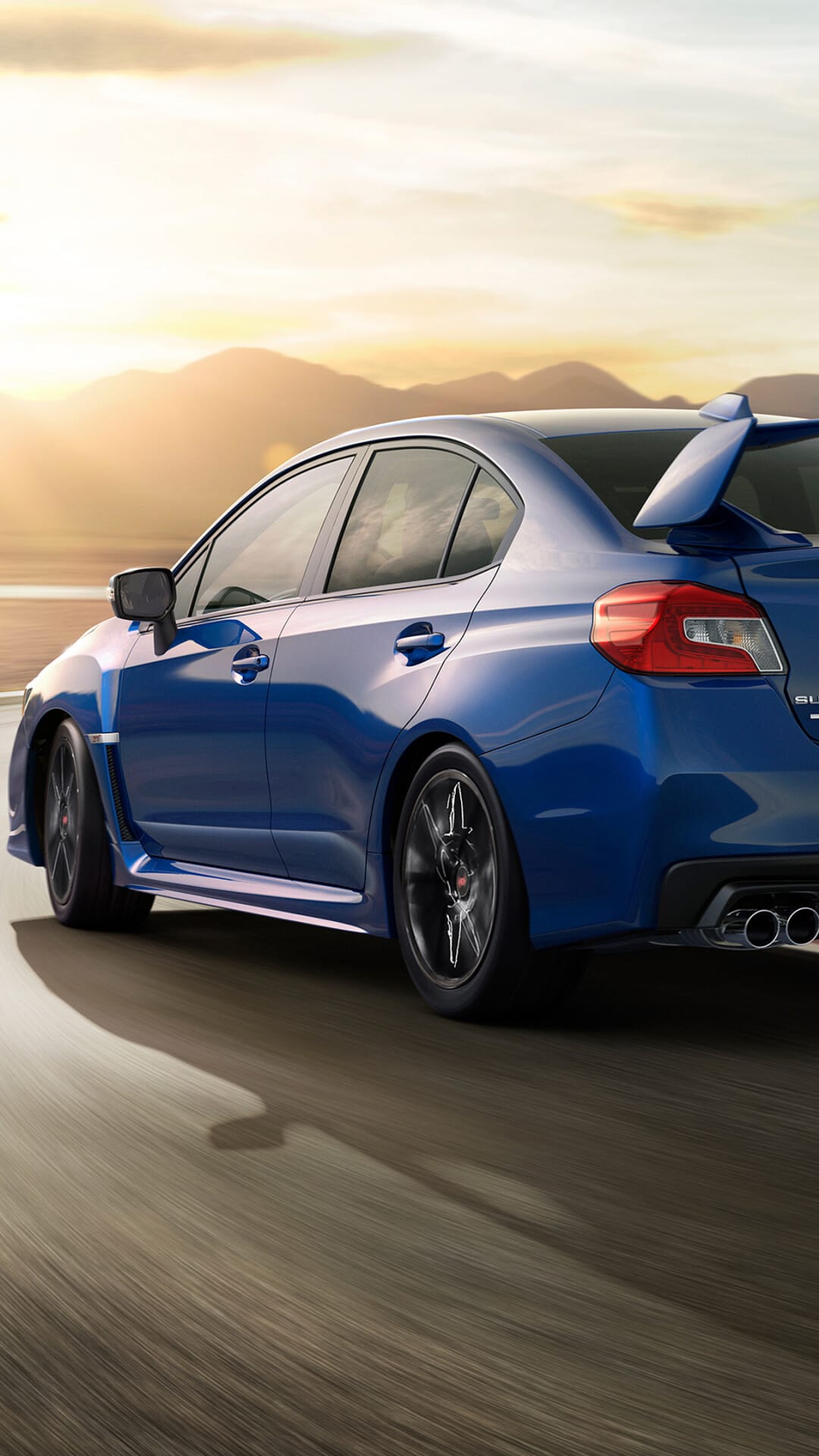 Blue sports sedan with a large rear wing driving on a curved road at sunset with mountain silhouettes.