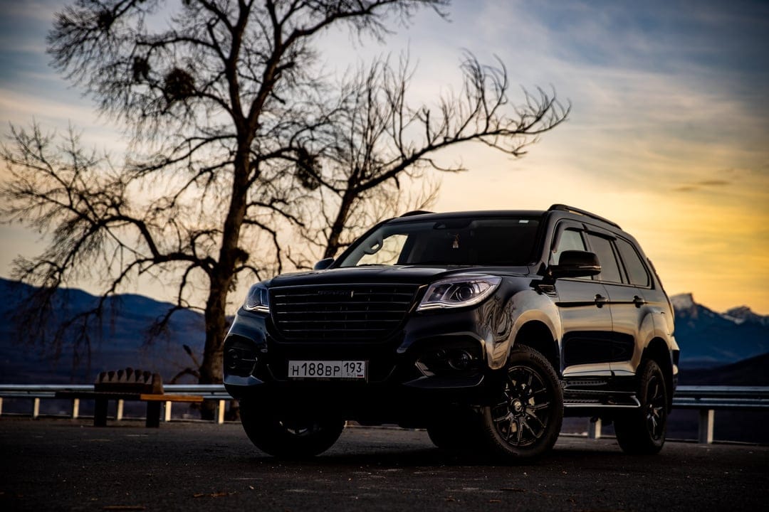 Black Haval H9 SUV parked on a mountain overlook at sunset with a large bare tree and blue peaks behind.