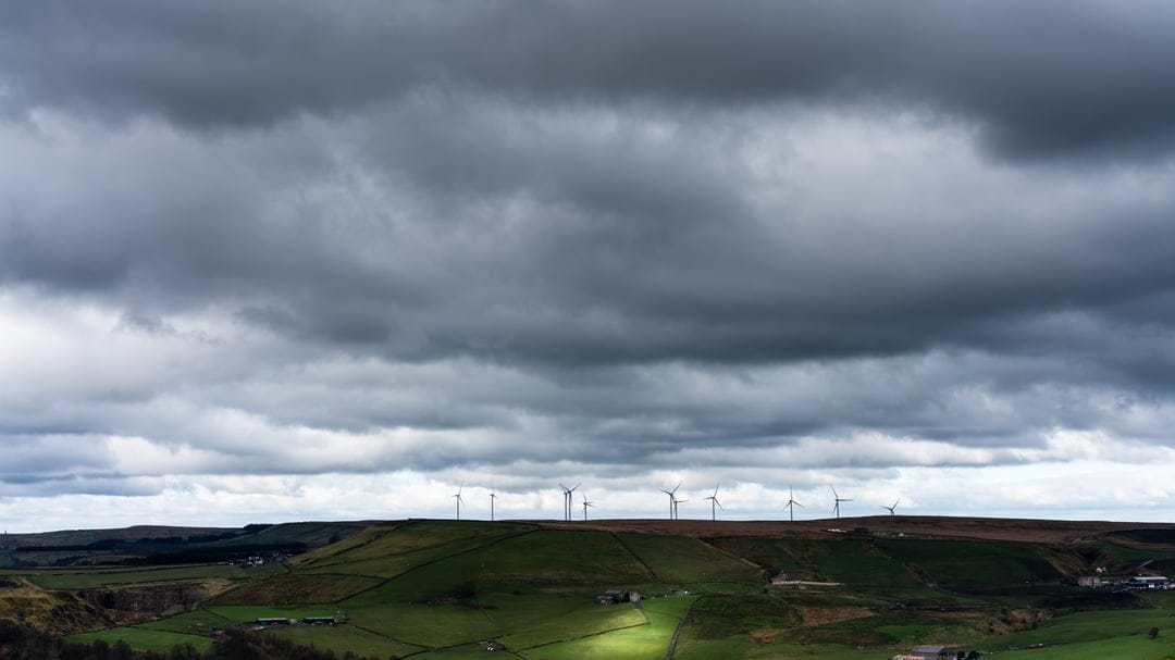 Wind turbines stand on rolling green hills beneath a dramatic dark sky with scattered patches of sunlight.