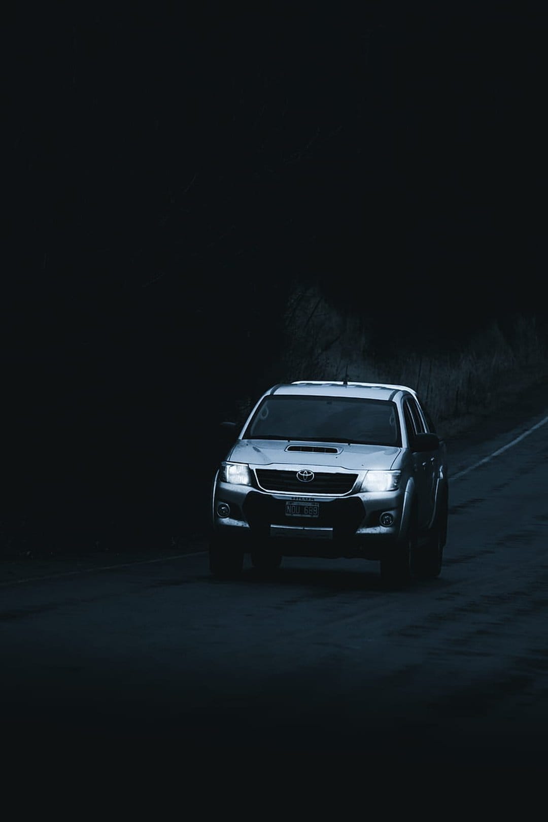 Silver pickup truck with glowing headlights driving down a dark road through a wooded area during the night.
