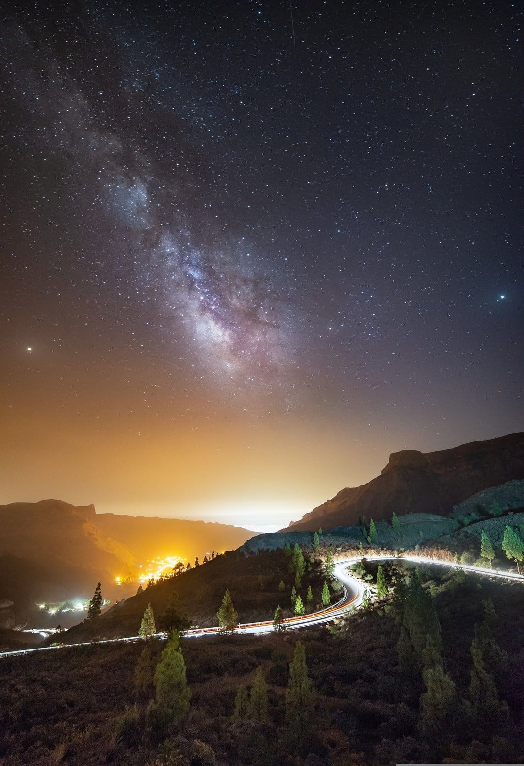 The Milky Way galaxy arches over a winding mountain road with car light trails and a glowing distant town.