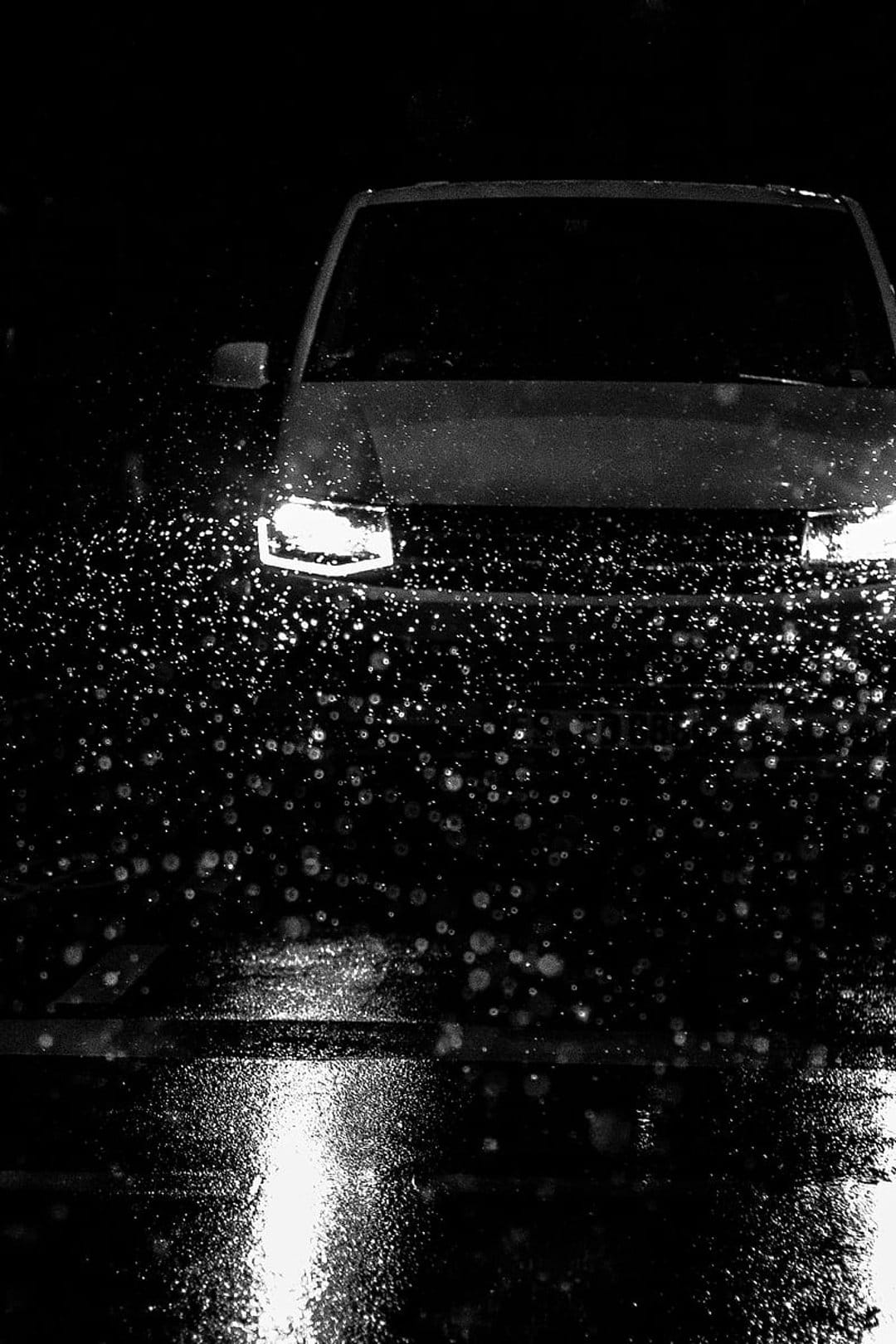 Car headlights pierce through night rain, illuminating water splashes on a dark, wet road in high contrast.