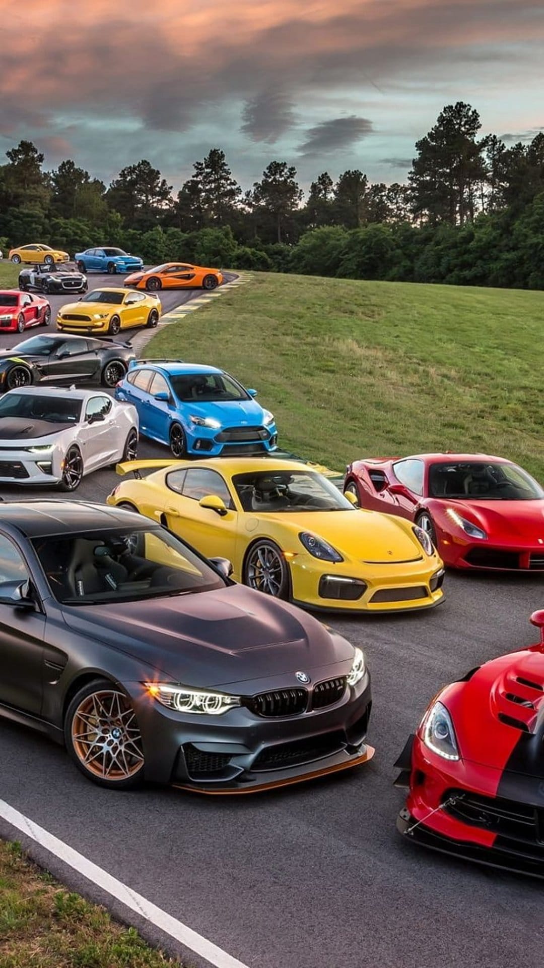 Exotic supercars lined up on a professional race track asphalt under a dramatic orange and purple sunset sky.