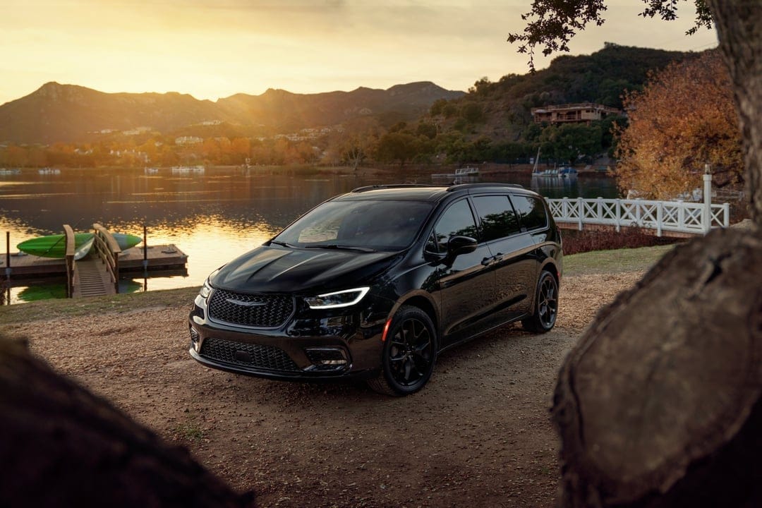 Black minivan on a dirt path by a lake at sunset with mountains and a wooden pier in the background.