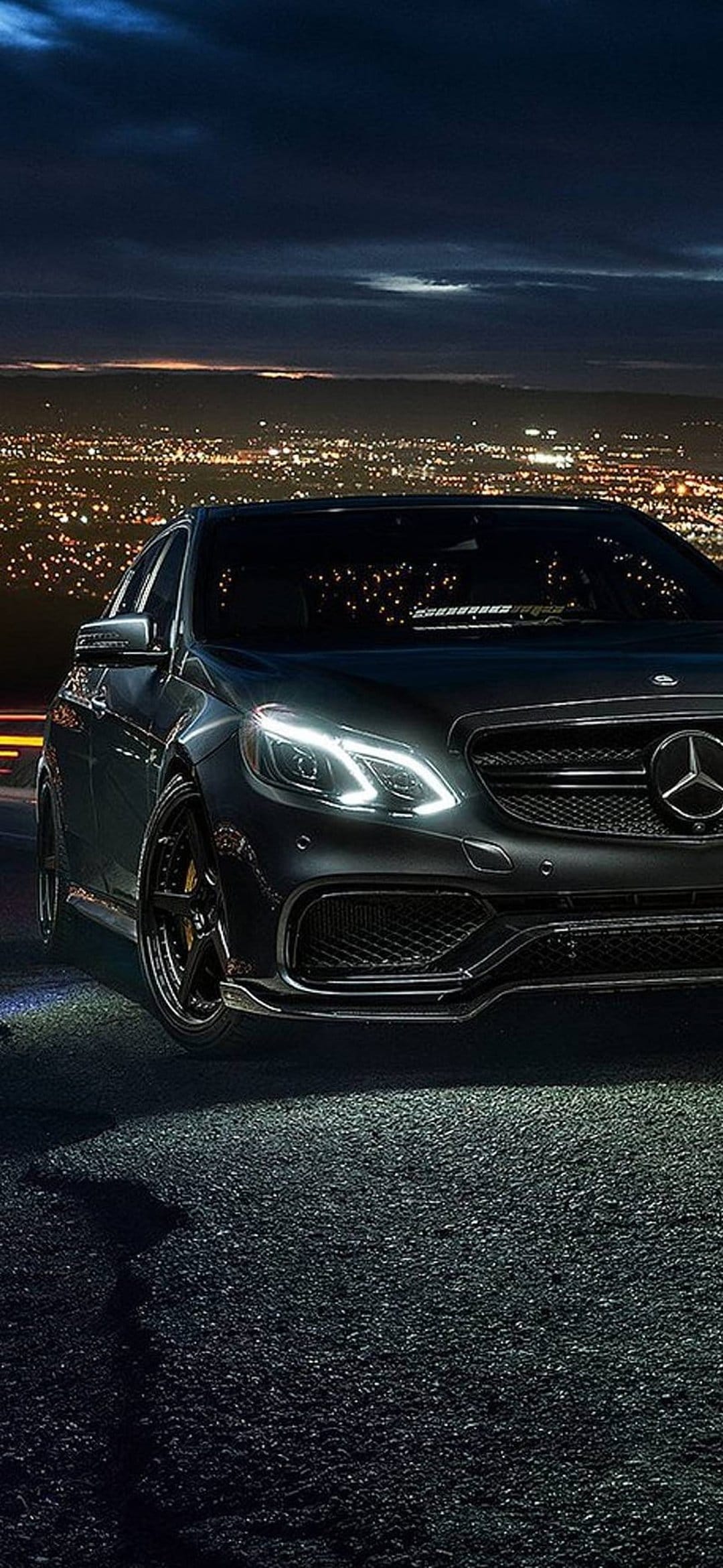 Black Mercedes-Benz E-Class sedan parked on asphalt with glowing headlights and a twinkling city backdrop.