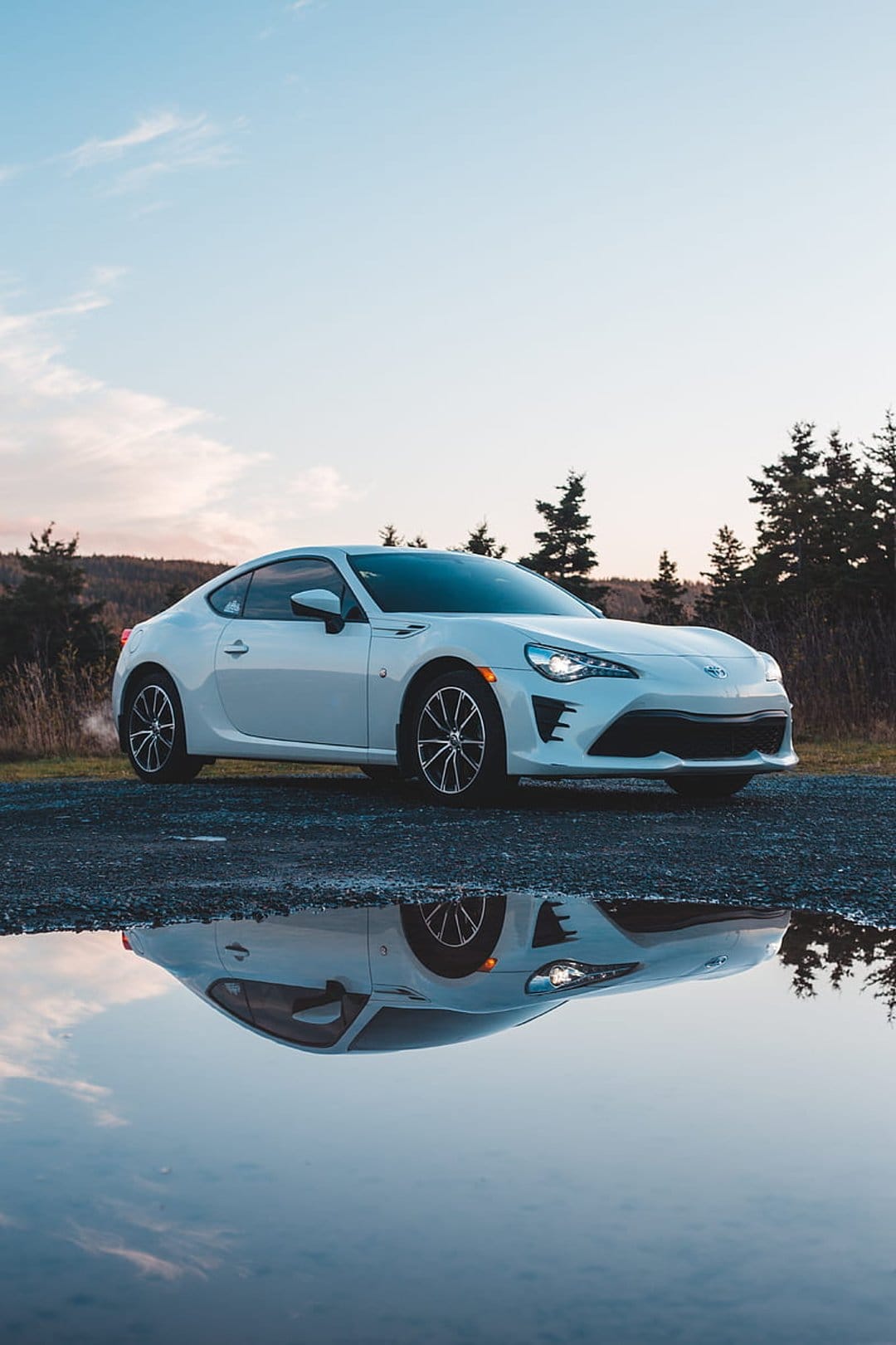 White Toyota 86 sports car reflects in a ground puddle during a colorful sunset amidst towering pine trees.