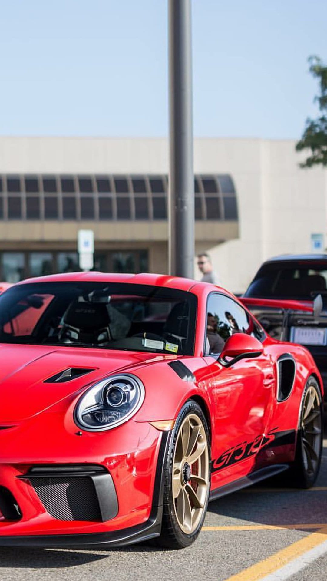 Red Porsche 911 GT3 RS with gold wheels parked on asphalt against a backdrop of trees and a modern building.