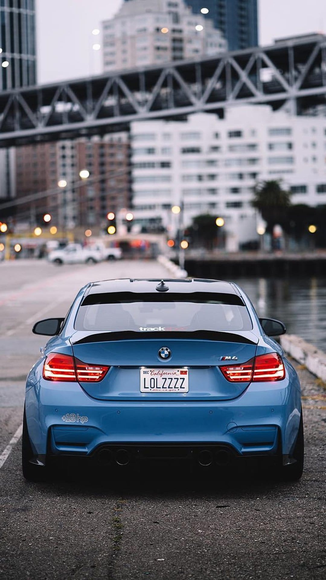 Blue BMW M4 rear view parked on a coastal road at dusk with a city skyline and bridge in the background.