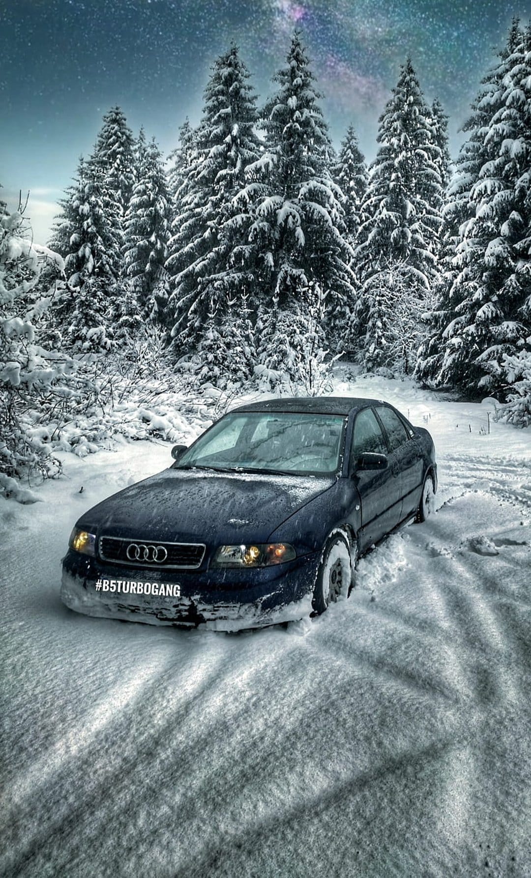 Dark blue Audi B5 sedan partially covered in snow on a forest road under a starry night sky during winter.
