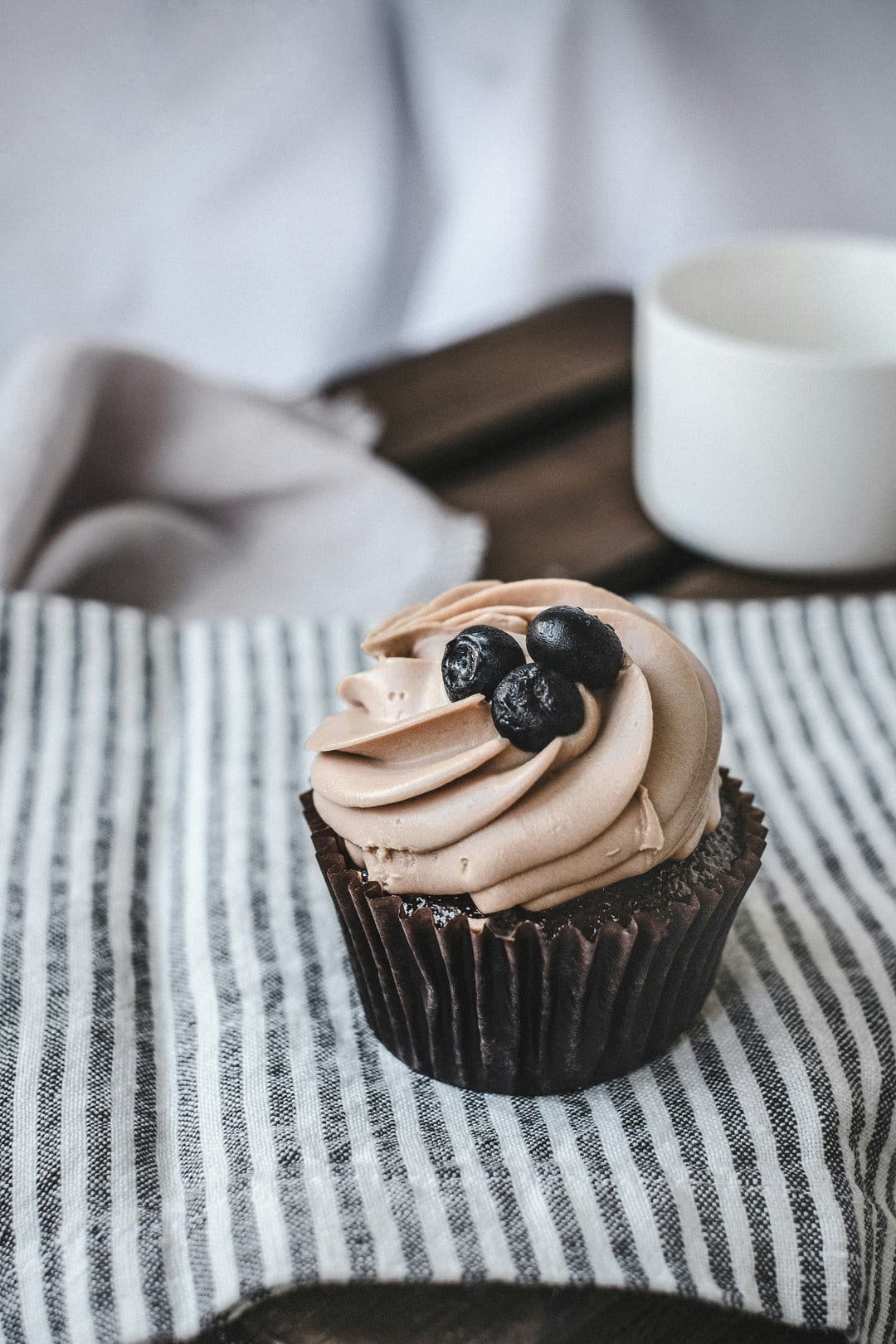 Chocolate cupcake topped with swirls of frosting and blueberries sits on a striped napkin over a wooden table.