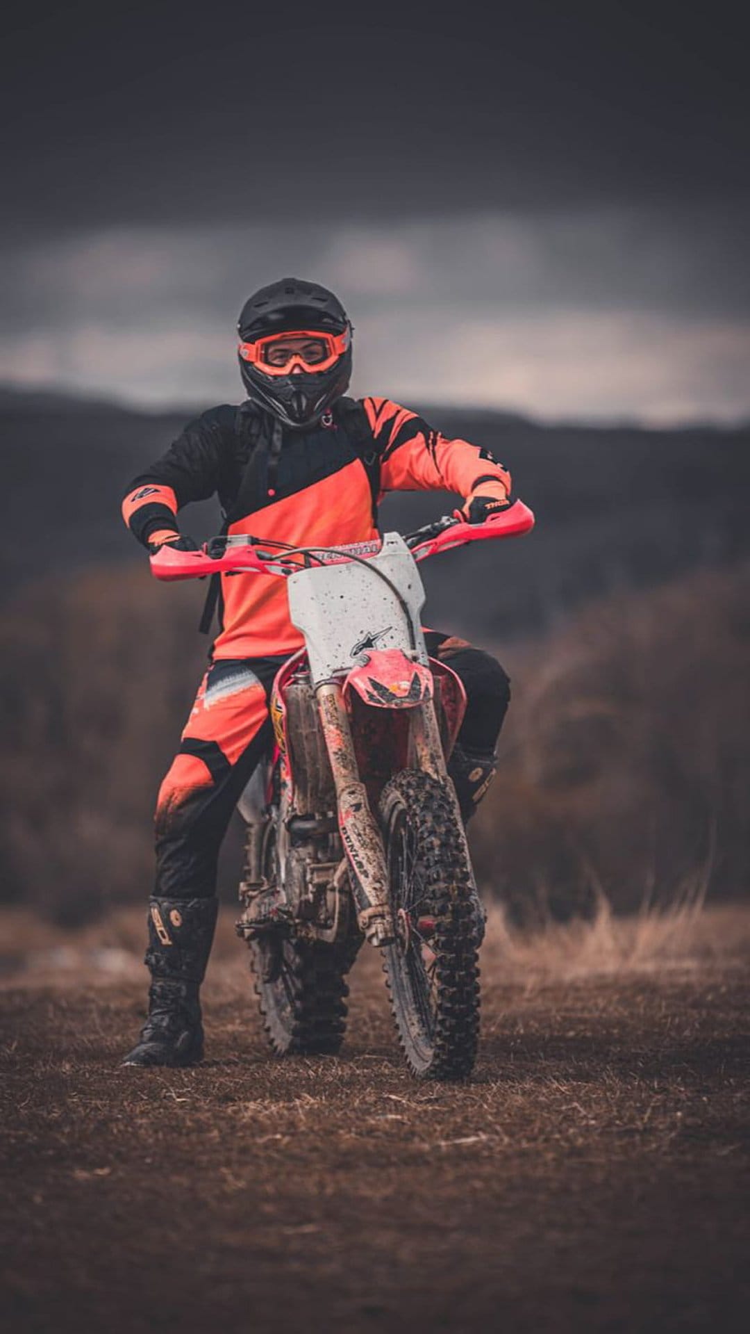Motocross racer in bright gear on a muddy motorcycle sits before a vast landscape of rolling hills and clouds.