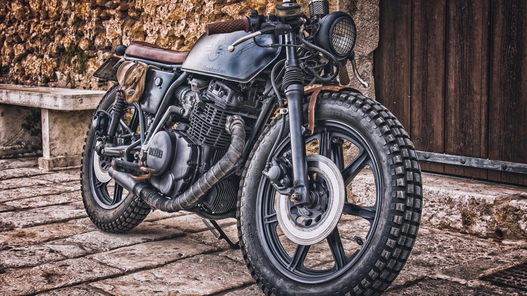 Custom black Yamaha motorcycle with knobby tires and brown leather seat parked against an old stone wall.