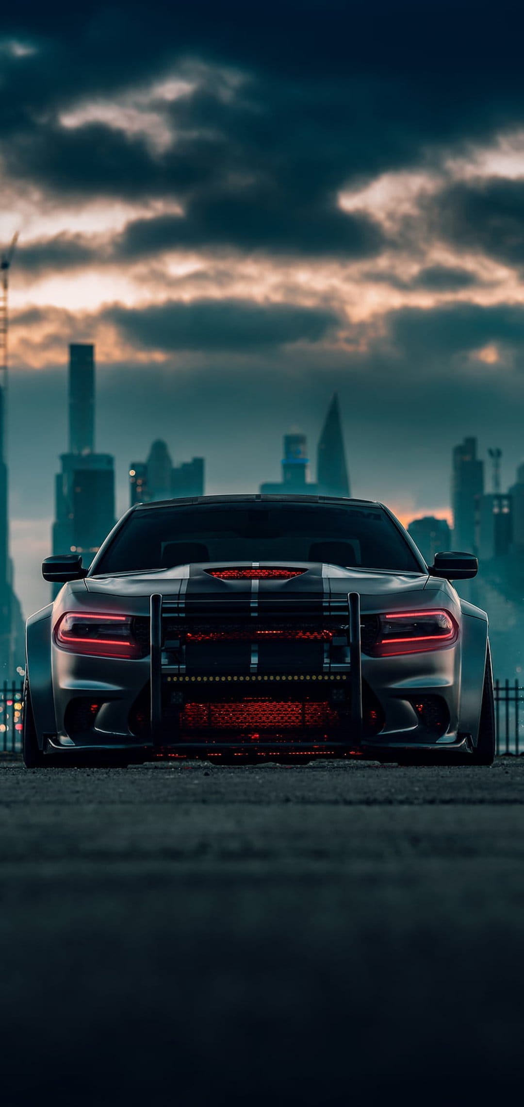 Dodge Charger with red LED headlights and a black bull bar parked in front of a cloudy city skyline at night.