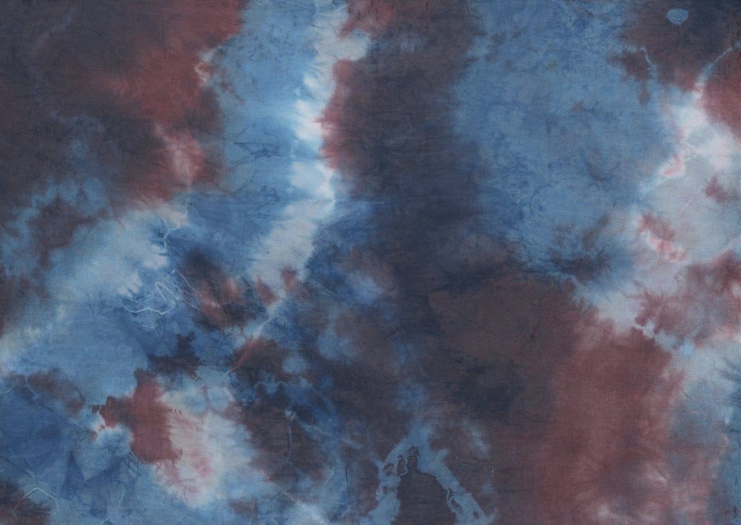 Blue and maroon tie-dye fabric swirls with marbled white textures creating a fluid and organic abstract pattern.