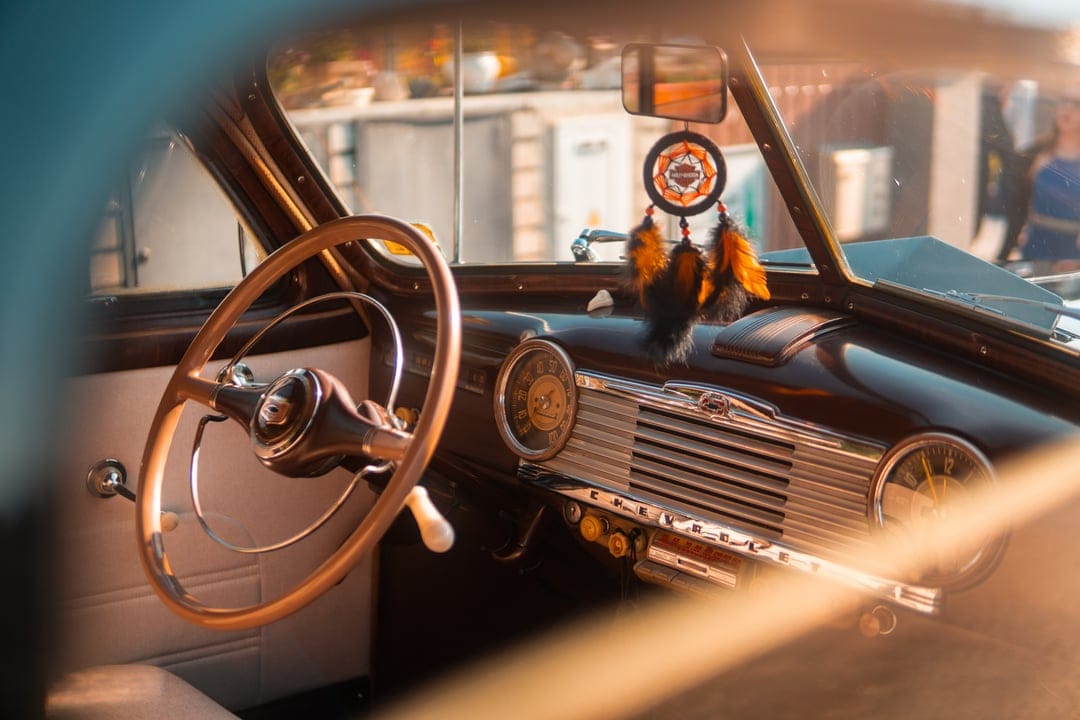 Wooden steering wheel and vintage car dashboard with chrome gauges and a dreamcatcher in warm lighting.