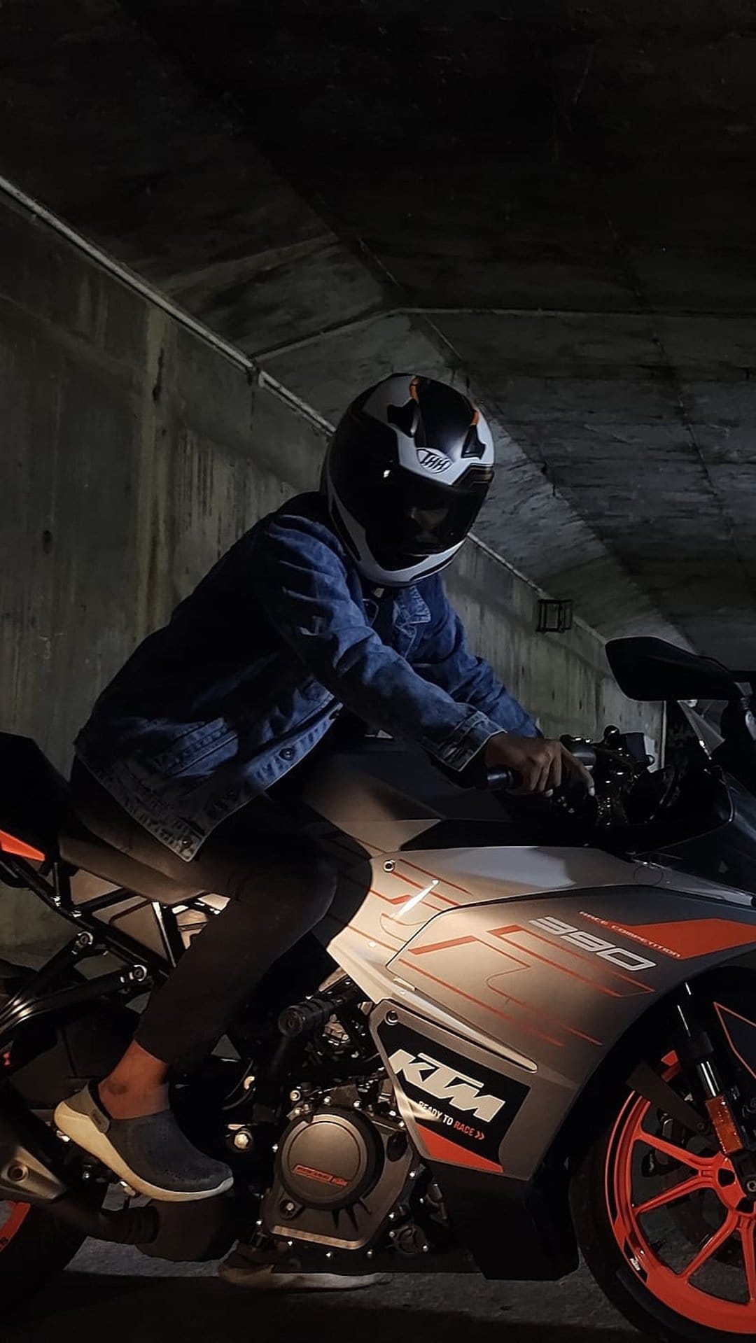 Motorcyclist in a denim jacket sits on a KTM 390 Duke with orange wheels inside a dark industrial tunnel.