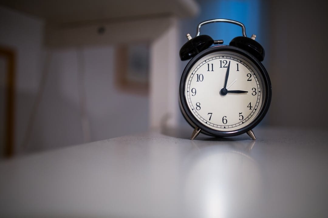 Black twin-bell alarm clock with silver metal accents sits on a white table against a blurred indoor background.