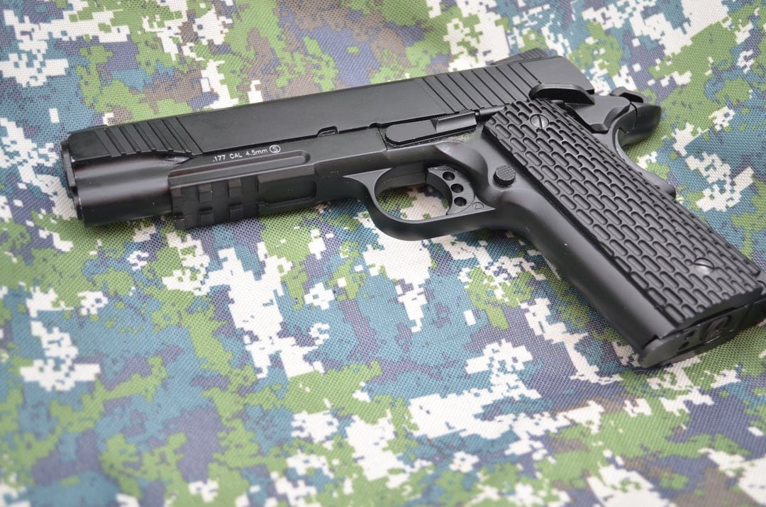 Black 1911 airsoft pistol with textured grips lies on green and blue camouflage fabric in a detailed close-up.