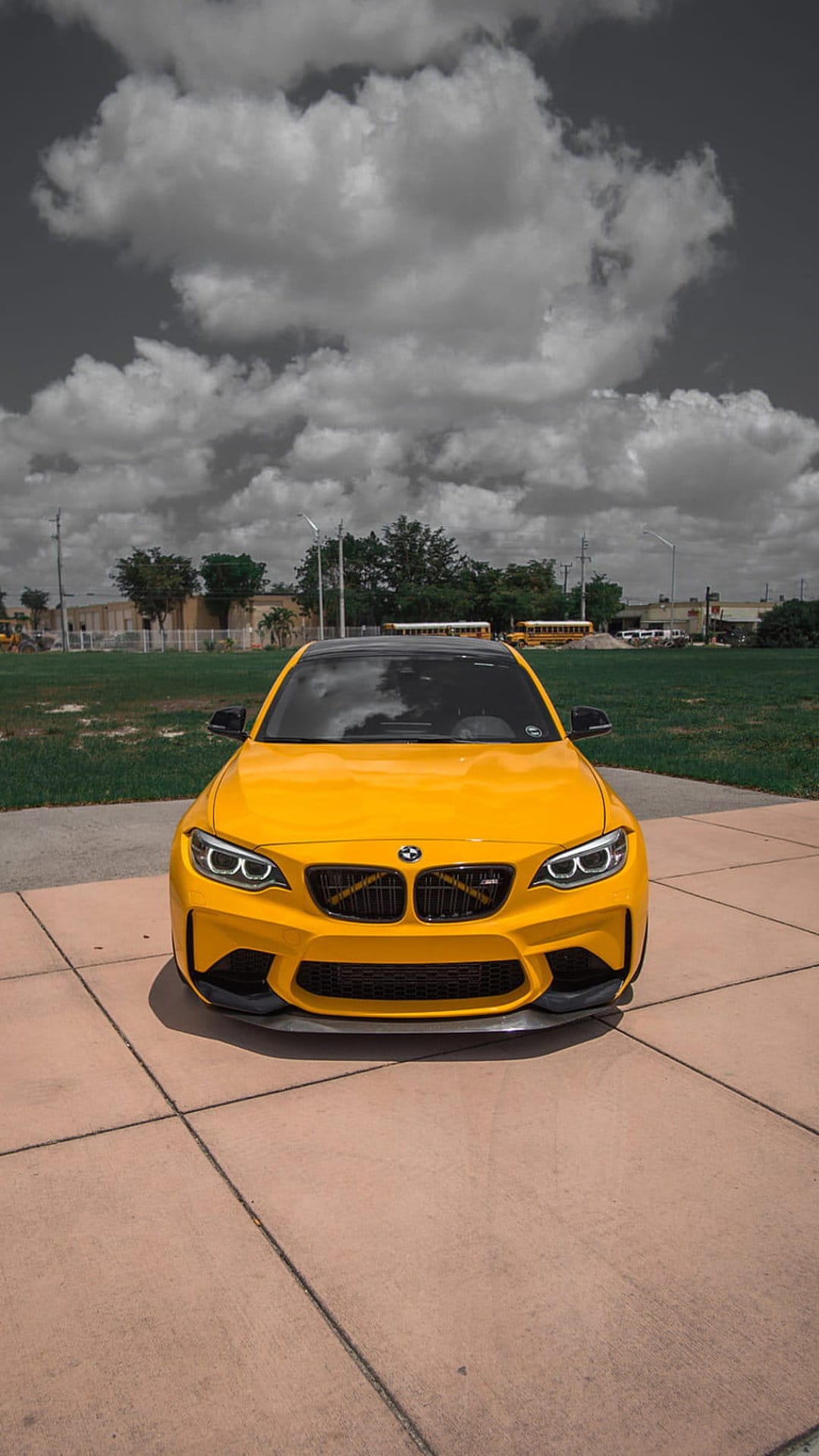 Yellow BMW M2 sports car with a black roof parked on concrete under a dark, textured cloudy sky near a field.