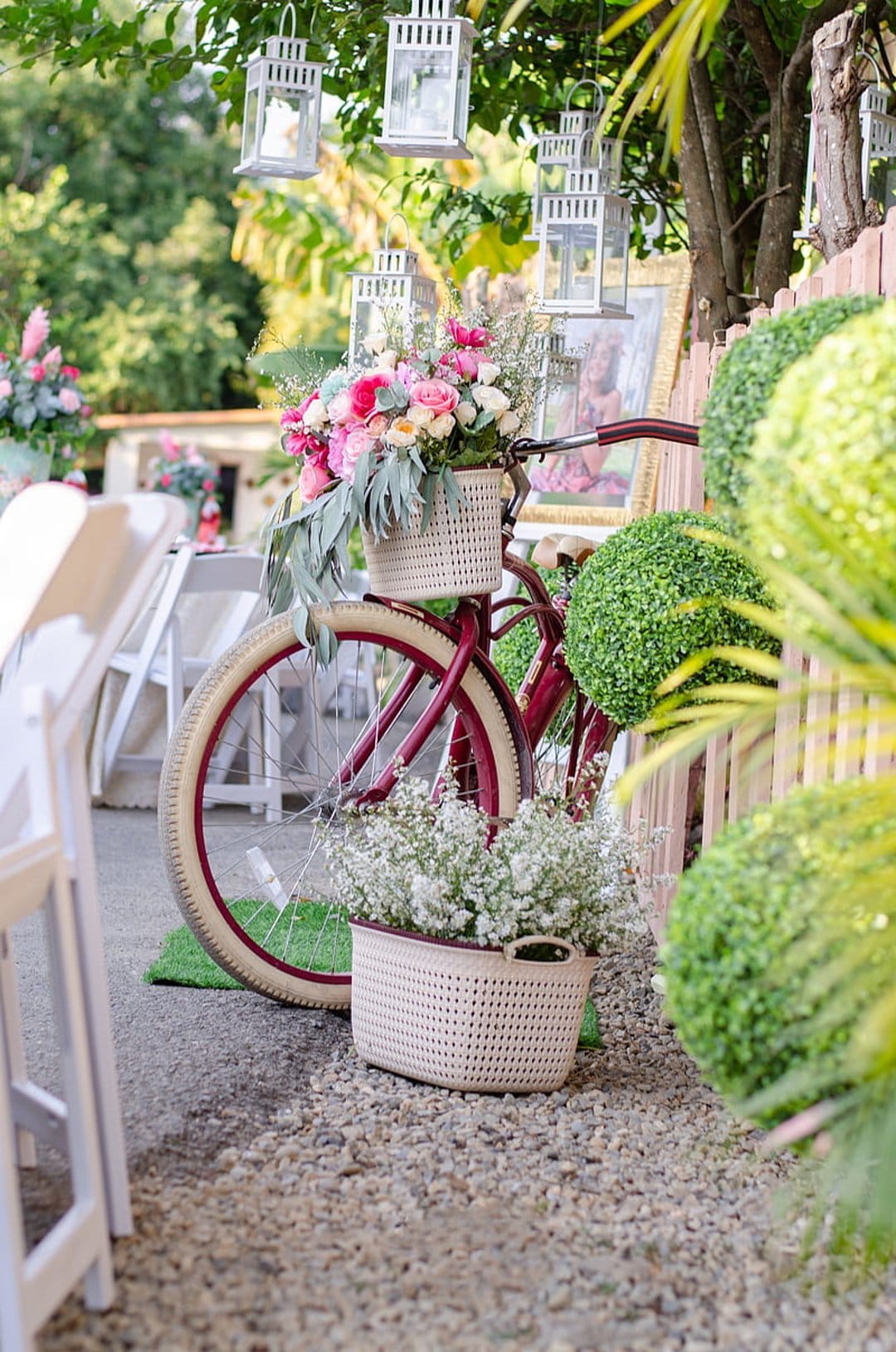 Decorative bicycle with a flower-filled basket stands near hanging lanterns and lush garden greenery at dusk.