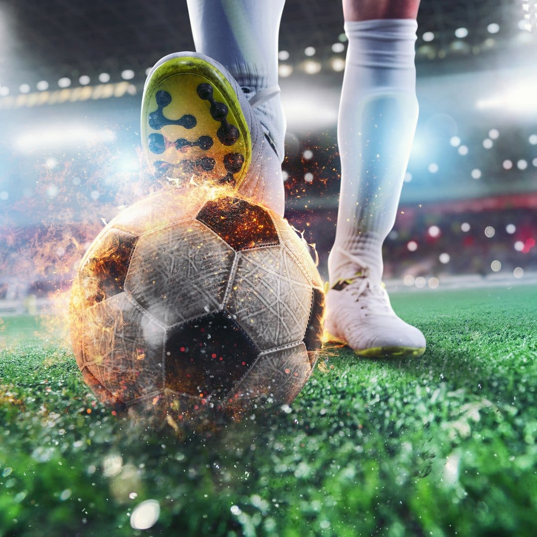 Soccer ball engulfed in orange flames being kicked by a player's foot in a stadium under bright floodlights.
