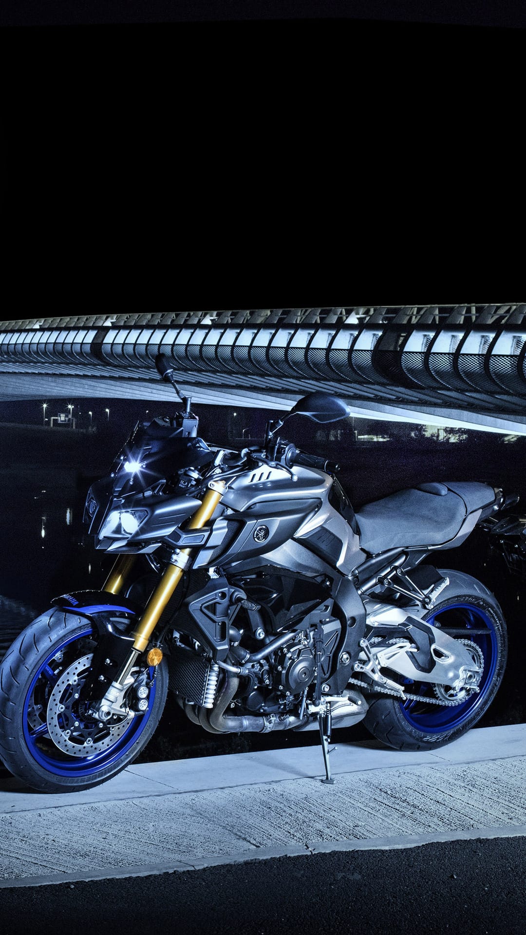 Yamaha MT-10 motorcycle with blue wheels parked beneath a modern bridge structure with glowing city lights.