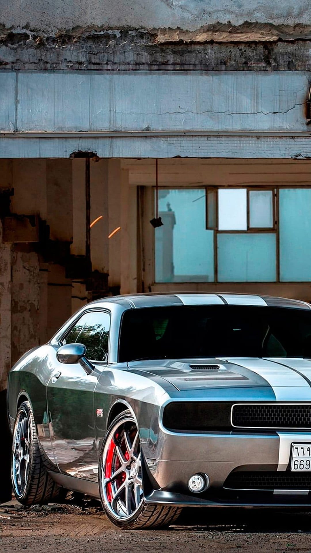 Silver Dodge Challenger with racing stripes parked by a weathered concrete wall in an urban industrial zone.